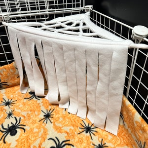 May include: White felt spider web hammock with fringe hanging from a white wire cage with a black background. The cage has a black and orange spider print fleece bedding.