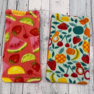 May include: Two rectangular fleece pads featuring fruit patterns. The left pad has a coral background with strawberries, watermelon slices, cherries, and lime wedges. The right pad has a light green background with various fruits.