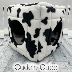 May include: A soft, plush cube-shaped pet bed featuring a black and white cow print pattern. The structure is constructed from quilted fabric panels and includes large, circular openings on the sides. The text Cuddle Cube is visible below.