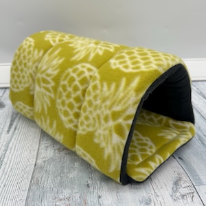May include: A yellow and white pineapple-patterned fleece tunnel bed for small pets. The interior is black. The bed is designed with a rounded shape and is made of soft material. The tunnel is designed to provide a cozy and secure space for small animals.