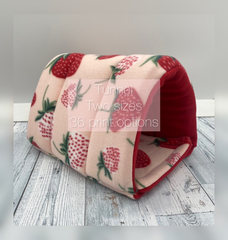 May include: A soft, arched pet tunnel featuring a light pink fleece exterior with a repeating red strawberry print and green leaves. The interior is lined with solid red fabric. Text on the image reads: Tunnel, Two sizes, 36 print options.
