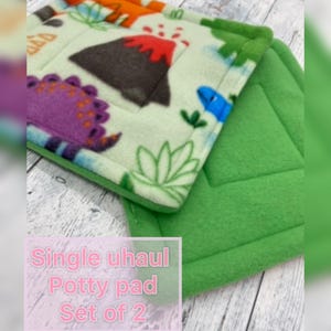 May include: A set of two square potty pads. One pad features a light green fleece fabric with a colorful dinosaur and volcano print. The second pad is solid green fleece. The text on the image reads: Single uhaul Potty pad Set of 2.