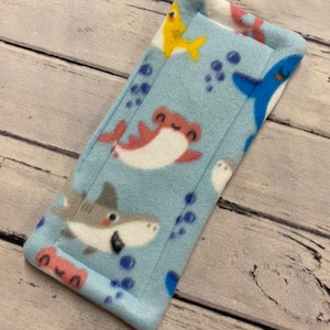 May include: A rectangular fleece fabric strip featuring a light blue background with a repeating pattern of various sharks in shades of gray, red, and yellow, accompanied by small dark blue bubbles scattered across the soft, plush material.