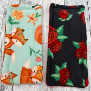 May include: Two rectangular fabric pads with rounded corners and stitched edges. The left pad features a light mint green background with orange foxes and yellow flowers. The right pad has a black background with red roses and green leaves.