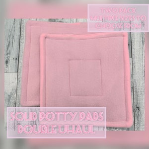May include: Two pink solid potty pads, double Uhaul, with a square design in the center. The pads are a soft pink color and have a stitched border. The text "TWO PACK MULTIPLE SIZES TO CHOOSE FROM" is in the upper right corner.