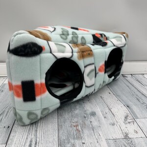 May include: A soft, light mint green fleece pet tunnel featuring a repeating sushi print with orange, black, and white details. The tunnel has two circular openings with black fabric trim, providing a cozy space for small animals to crawl through.