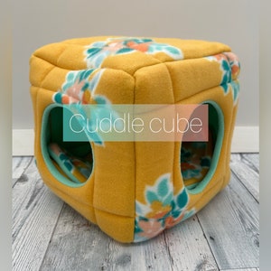 May include: A yellow fleece cuddle cube with floral accents in teal, peach, and white. The cube has three openings with teal trim and a floral patterned interior. The text "Cuddle cube" is displayed on the front.