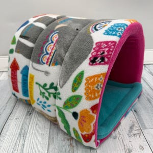 May include: A tunnel-shaped pet bed made of white fleece with a colorful pattern of gray elephants, green leaves, orange flowers, and red and blue arrows. The interior is lined with solid teal fabric, and the edges are trimmed in bright pink.