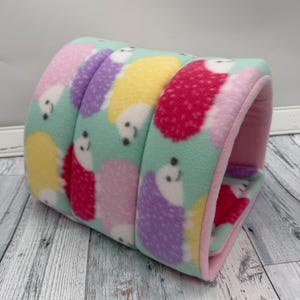 May include: A cylindrical fleece tunnel for small pets featuring a light mint green background with a repeating pattern of colorful hedgehogs in shades of yellow, purple, and red. The interior of the tunnel is lined with solid light pink fleece.
