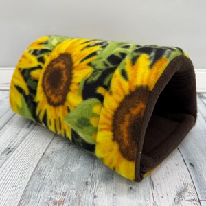 May include: A cylindrical pet tunnel made of soft fleece fabric featuring a vibrant sunflower pattern with large yellow petals and dark brown centers against a green and black background, finished with a solid dark brown fleece interior lining.