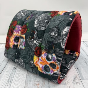 May include: A tunnel-shaped pet bed made of soft fleece fabric featuring a dark gray background with a repeating pattern of colorful sugar skulls and red roses. The interior of the tunnel is lined with a solid deep red fabric.