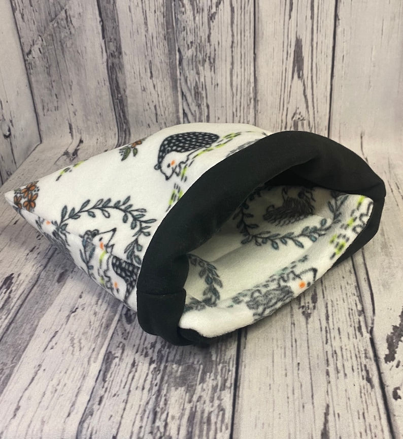 May include: A soft fabric pouch featuring a white base with a repeating pattern of hedgehogs, green leaves, and orange flowers. The opening of the pouch is finished with a wide, solid black fabric trim that folds over the top edge of the item.