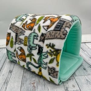 May include: A small pet bed with a white exterior patterned with orange foxes, green lizards, and brown trees. The interior is a solid light green color. The bed is made of a soft, plush material.
