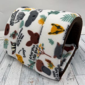 May include: A cylindrical pet tunnel made of white fleece fabric featuring a repeating pattern of Australian animals including dingoes, koalas, and platypuses, with dark brown interior lining and black text labels identifying each animal species.