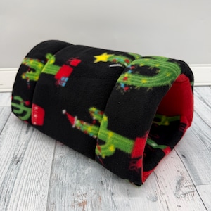 May include: A cylindrical fleece tunnel featuring a black background with a repeating pattern of green cacti wearing Santa hats and decorated with colorful lights. The interior of the tunnel is lined with a solid red fabric.