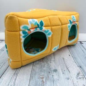 May include: A rectangular yellow fleece pet tunnel featuring a floral pattern in shades of teal, white, and orange. The structure has two circular openings with teal trim, a quilted exterior texture, and a soft interior lining visible through the holes.