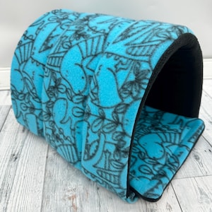 May include: A cylindrical pet tunnel made of bright blue fleece fabric featuring a black line-art pattern of various dinosaurs and foliage. The interior of the tunnel is lined with solid black fabric, and the structure has a soft, padded design.