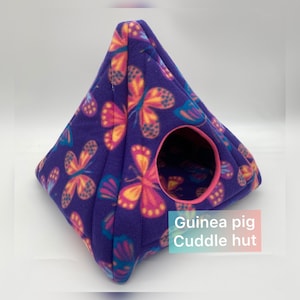 May include: A pyramid-shaped fleece cuddle hut for small pets featuring a purple background with a repeating pattern of orange, pink, and blue butterflies. The structure has a circular entrance trimmed in bright pink fabric and includes the text Guinea pig Cuddle hut.