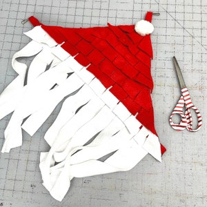 May include: A red and white fabric toy with fringe. The toy is shaped like a triangle and has a white pom-pom on top. A pair of red and white striped scissors are also in the image.