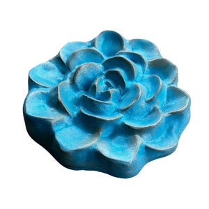 May include: A turquoise-colored, flower-shaped decorative object. The object has multiple layers of petals, with a textured surface and a slightly weathered appearance. The flower is a solid, three-dimensional piece, likely made of a durable material.