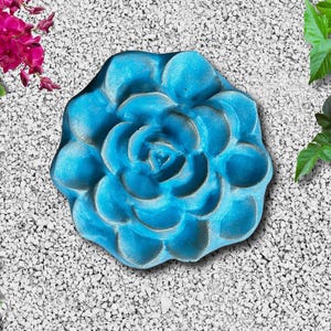 May include: A turquoise-colored, flower-shaped garden stepping stone. The stone has a detailed, textured surface with a central spiral design. The stone is surrounded by small white pebbles and greenery.