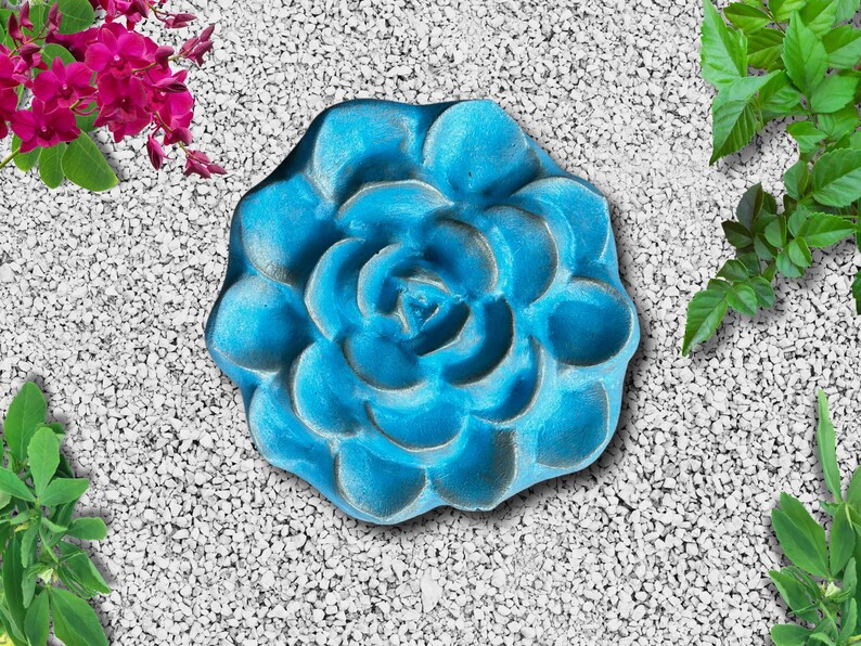 May include: A turquoise-colored, flower-shaped garden stepping stone. The stone has a detailed, textured surface with a central spiral design. The stone is surrounded by small white pebbles and greenery.