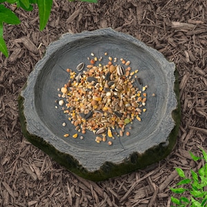 May include: A gray, tree-stump-shaped bird feeder filled with a mix of seeds, nuts, and grains. The feeder sits on a bed of brown mulch, with green foliage in the background. A natural and rustic design for outdoor use.