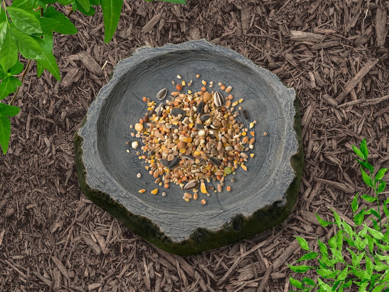 May include: A gray, tree-stump-shaped bird feeder filled with a mix of seeds, nuts, and grains. The feeder sits on a bed of brown mulch, with green foliage in the background. A natural and rustic design for outdoor use.