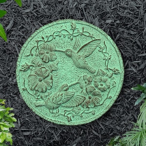 May include: A round, weathered-look, green stepping stone with a hummingbird and floral design. The stone features two hummingbirds and several flowers, with vines and leaves around the edge. The stone is set on a bed of black mulch.