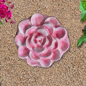 May include: A pink, rose-shaped stepping stone. The stone is surrounded by small, tan pebbles and greenery. The rose has a detailed, layered petal design and a light pink color with white highlights.