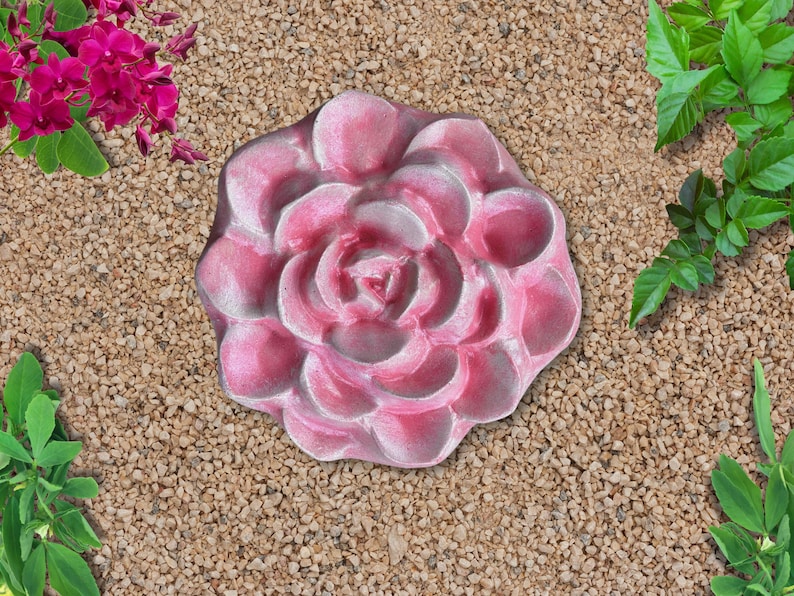 May include: A pink, rose-shaped stepping stone. The stone is surrounded by small, tan pebbles and greenery. The rose has a detailed, layered petal design and a light pink color with white highlights.