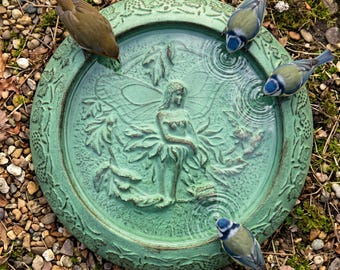 May include: A round, weathered green bird bath with a fairy design in the center. Four small birds are drinking from the water-filled basin. The bird bath is surrounded by small pebbles and greenery.