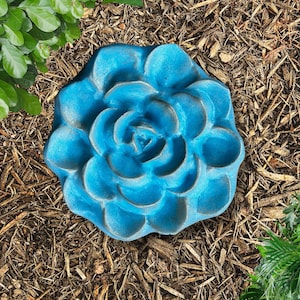 May include: A blue, flower-shaped garden stepping stone. The stone has a textured surface and is surrounded by mulch and greenery. The flower design is detailed with multiple petals and a central spiral.