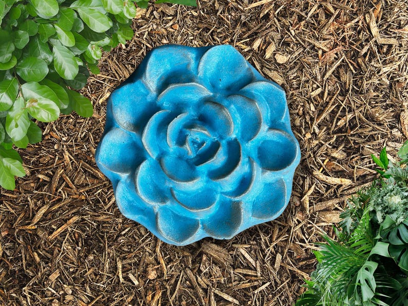May include: A blue, flower-shaped garden stepping stone. The stone has a textured surface and is surrounded by mulch and greenery. The flower design is detailed with multiple petals and a central spiral.