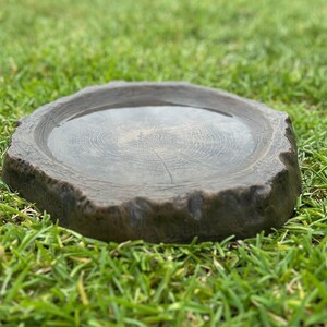 May include: A round, gray bird bath made of stone, resting on a bed of green grass. The bird bath has a textured surface and a shallow basin filled with water. The edges are rough and uneven, resembling a tree stump.