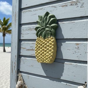 May include: A yellow and green pineapple-shaped wall decoration. The pineapple is mounted on a weathered, light blue wooden surface. A decorative net with seashells hangs below. A beach scene is visible in the background.