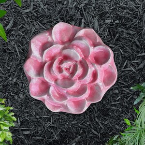 May include: A pink, flower-shaped stepping stone with a detailed petal design. The stone is set against a backdrop of black mulch and green foliage, creating a garden-inspired aesthetic.
