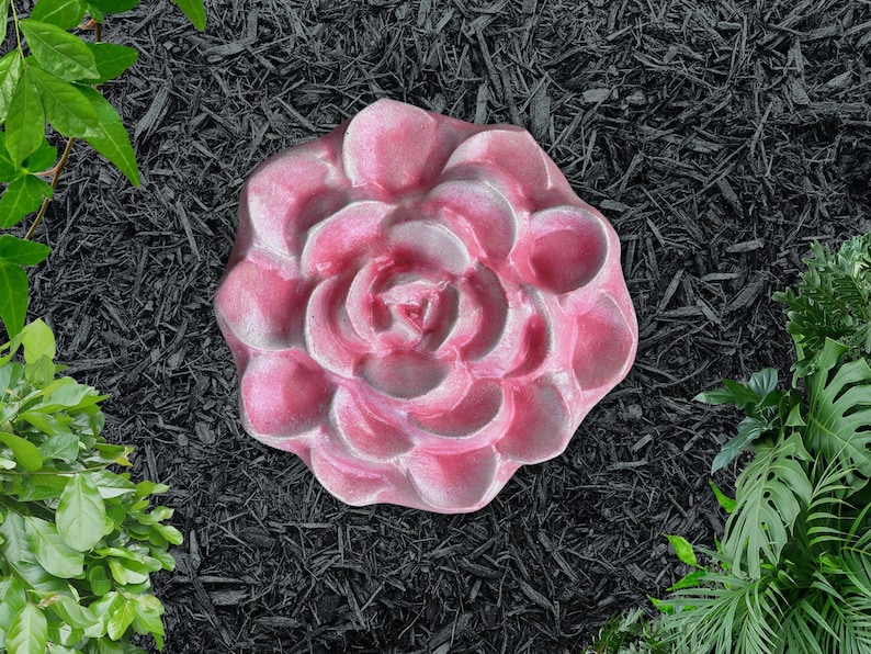 May include: A pink, flower-shaped stepping stone with a detailed petal design. The stone is set against a backdrop of black mulch and green foliage, creating a garden-inspired aesthetic.
