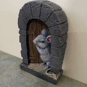 Op de afbeelding: Een grijs muisbeeldje dat uit een kleine stenen boog met een houten deur gluurt. De muis houdt zijn poot bij zijn mond en heeft roze oren.