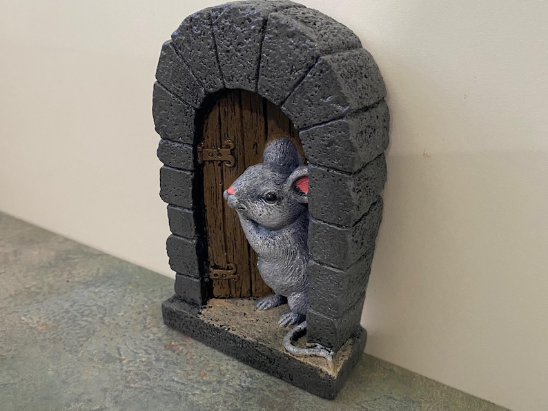 Op de afbeelding: Een grijs muisbeeldje dat uit een kleine stenen boog met een houten deur gluurt. De muis houdt zijn poot bij zijn mond en heeft roze oren.