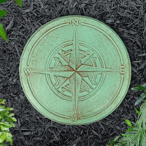 May include: A green compass-shaped stepping stone with a rusty brown compass design. The stone has the letters N, S, E, and W around the compass.