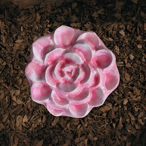 May include: A pink, flower-shaped decorative item with white accents. The item is made of a solid material and is set against a backdrop of brown mulch. The flower has multiple petals and a detailed center.
