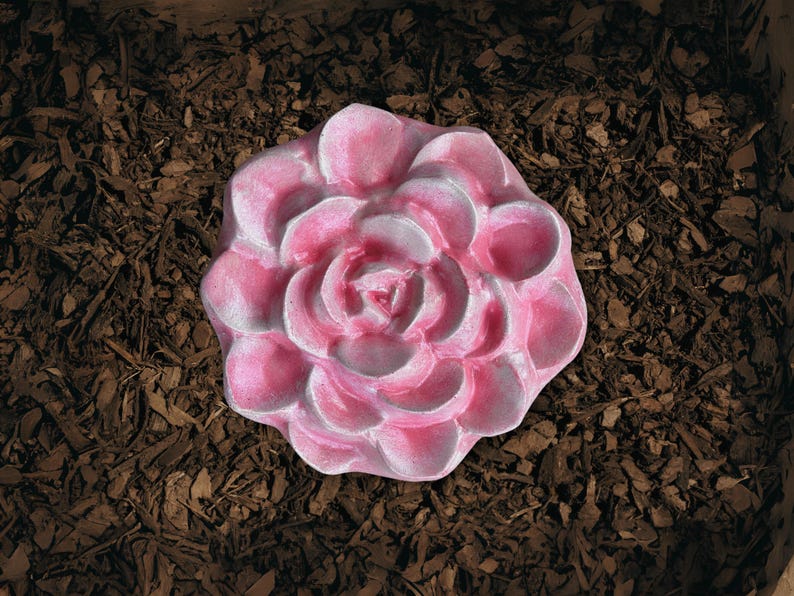 May include: A pink, flower-shaped decorative item with white accents. The item is made of a solid material and is set against a backdrop of brown mulch. The flower has multiple petals and a detailed center.