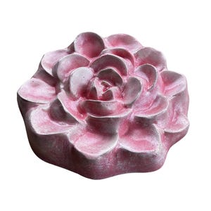 May include: A pink, flower-shaped decorative object. The object has multiple layers of petals, with a textured surface. The color is a soft pink with some areas of a lighter shade. The object is likely made of a solid material.