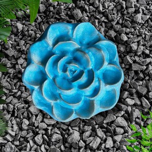 May include: A vibrant blue, flower-shaped decorative object. The object is made of a textured material and is surrounded by gray rocks and green foliage. The flower has multiple layers of petals and a detailed center.