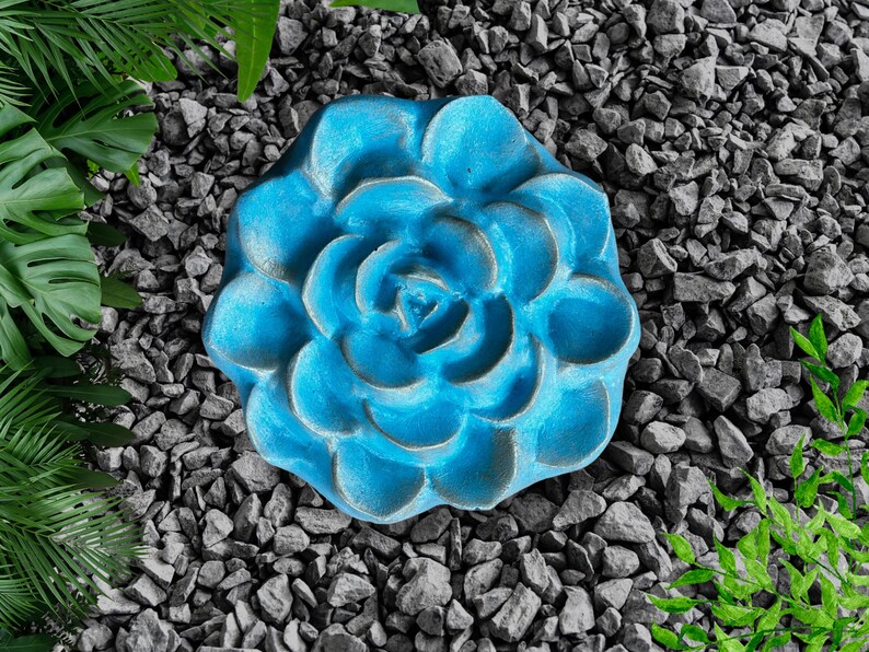May include: A vibrant blue, flower-shaped decorative object. The object is made of a textured material and is surrounded by gray rocks and green foliage. The flower has multiple layers of petals and a detailed center.