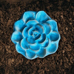 May include: A vibrant blue, flower-shaped garden ornament. The succulent-like design is set against a backdrop of brown mulch. The ornament's petals are detailed, creating a realistic appearance. This decorative piece adds a pop of color to any garden or outdoor space.