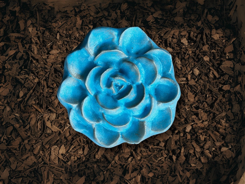 May include: A vibrant blue, flower-shaped garden ornament. The succulent-like design is set against a backdrop of brown mulch. The ornament's petals are detailed, creating a realistic appearance. This decorative piece adds a pop of color to any garden or outdoor space.