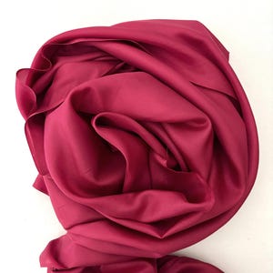 May include: A solid deep magenta fabric draped in soft, voluminous folds. The material has a smooth, matte texture with subtle shadows created by the overlapping layers of the cloth, forming a circular, rose-like arrangement of fabric.