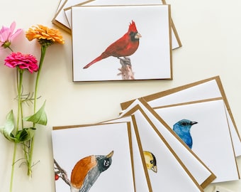 May include: A set of eight blank greeting cards featuring watercolor illustrations of different birds. The cards are brown on the outside and white on the inside. The birds include a cardinal, a bluebird, a robin, and a yellow bird.